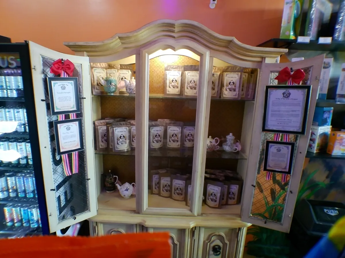 Cleo Baby tea bags and decorative teapots in a cream-colored cabinet at Jubilee, with framed tea descriptions on its open doors.